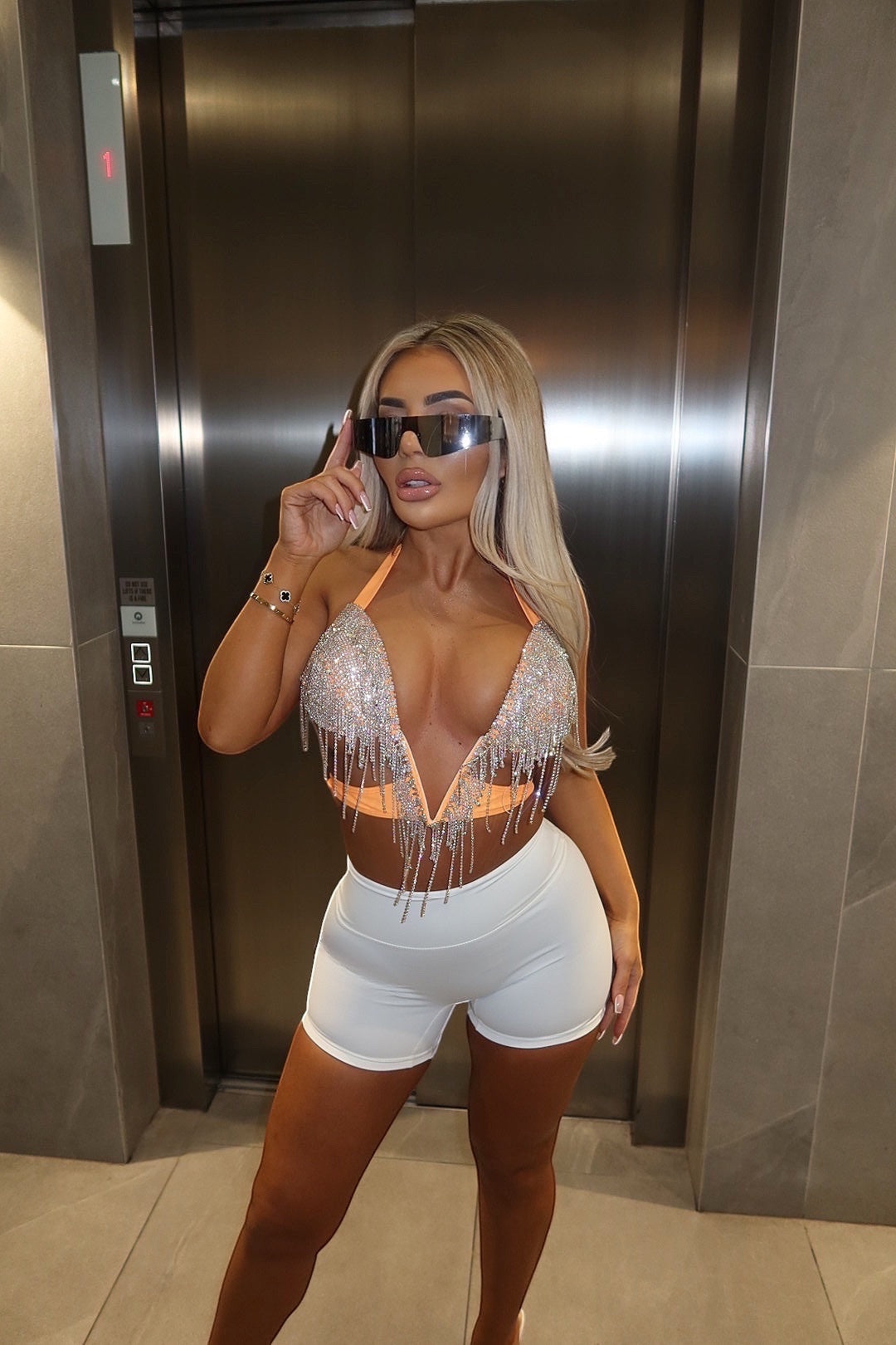 GLAMORZ coral diamond-embellished bra top with fringe detail worn by a blonde model with oversized sunglasses